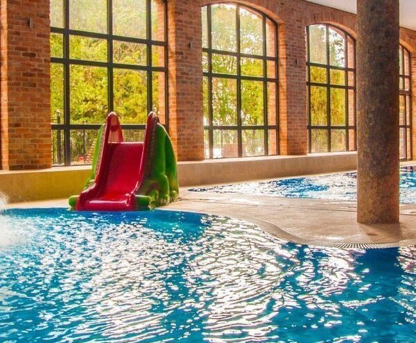 Bright indoor hotel pool with mushroom waterfall, two colorful children's slides, arched brick windows and wooden mezzanine