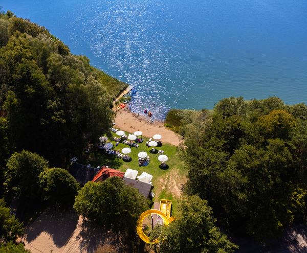 Private lakeside beach with white umbrellas and sun loungers on lawn, kayaks and boats at the shore, bright yellow waterslide.