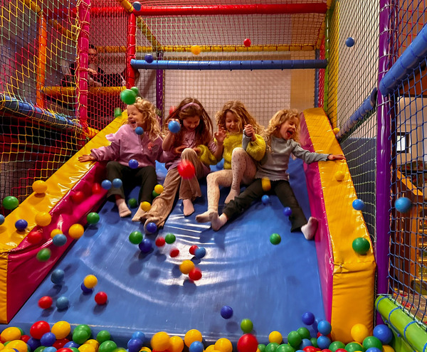 Family-friendly indoor play area: four laughing children sliding into a colorful ball pit on a padded soft-play slide.