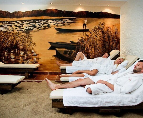 Hotel spa relaxation room with sand floor, cushioned loungers and three guests in white robes resting under a warm lakeside mural.