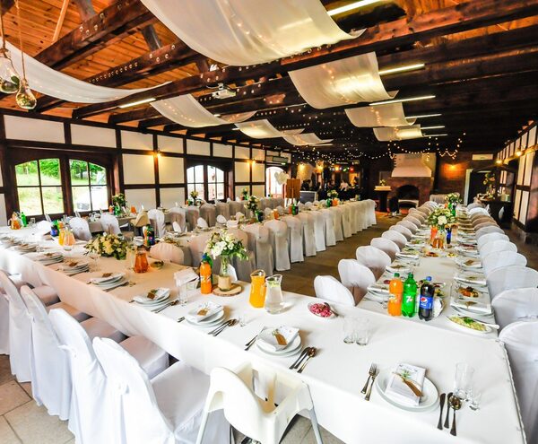 Rustic banquet hall with wooden beams, long white tables set for guests, floral centerpieces, draped ceiling fabric and fairy lights