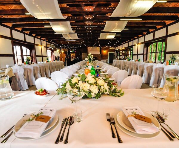 Rustic banquet hall set for a wedding with long white-linen tables, floral centerpieces, draped fabric and string lights.