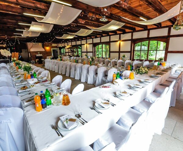 Rustic timber banquet hall for wedding with long linen-covered tables, white chair covers, floral centers, draped fabric, fairy lights, garden view.