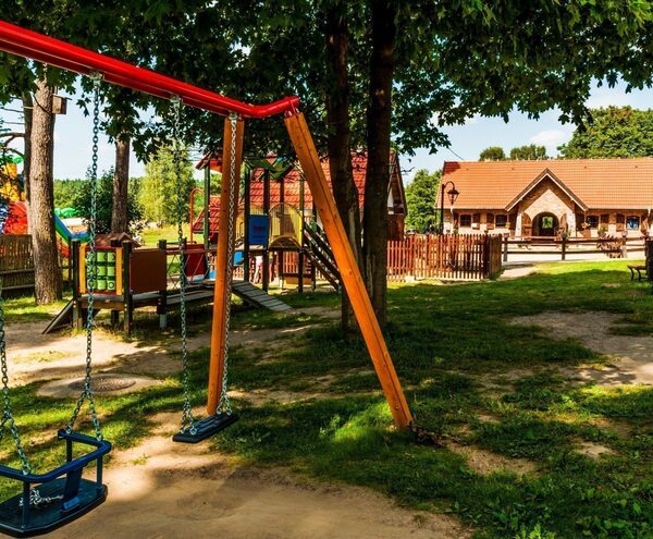 Shaded playground with swings, climbing structure and benches on a grassy hotel courtyard by a charming brick building.