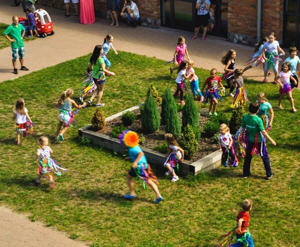 Children running and playing in a hotel courtyard garden, wearing colorful ribbon skirts with staff in green shirts supervising.