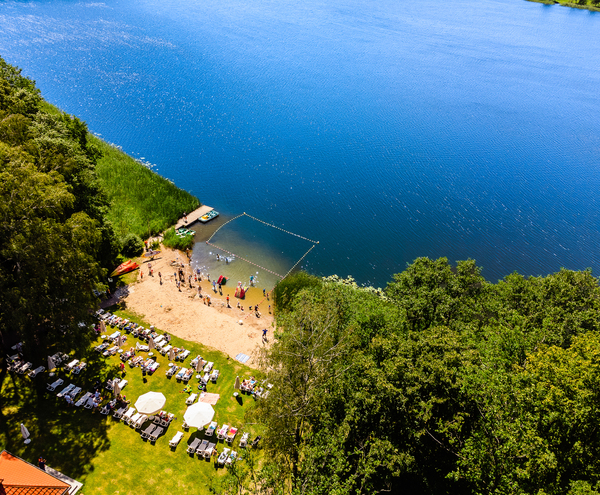 Lakeside beach with grassy sunbathing area, rows of loungers and umbrellas, sandy shore with swimmers, a dock with paddleboats and roped swim zone