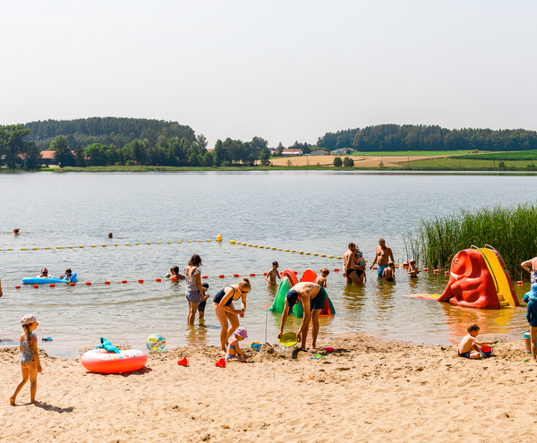 Family-friendly sandy lake beach with buoyed swim zone, kids playing on inflatable slide and shallow water, pastoral views.