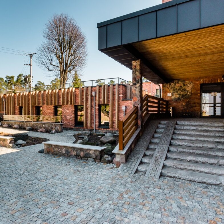 Covered hotel entrance with wooden ceiling, stone steps and ramp, brick façade, paved courtyard and landscaped rock beds.