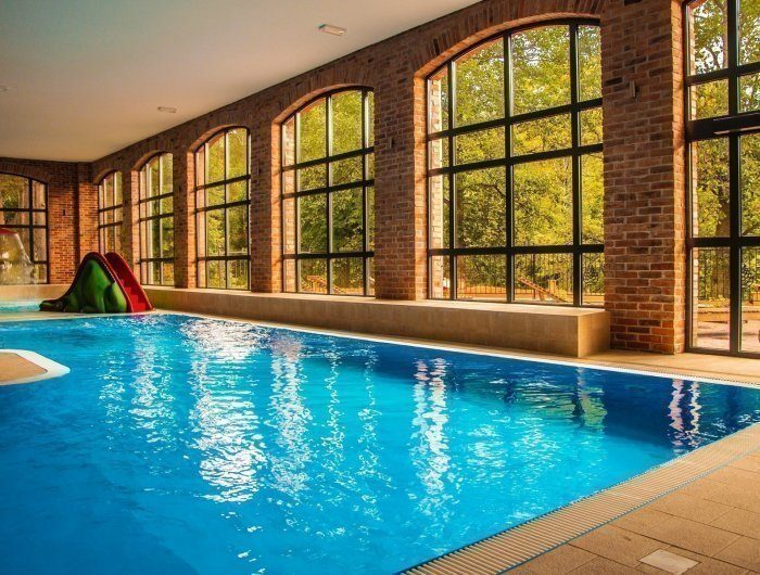 Indoor heated pool with arched brick windows, natural light, bench seating and a small children's slide overlooking trees.