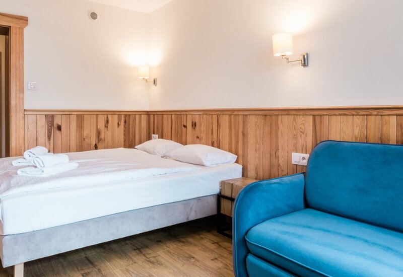 Przytulny pokój dwuosobowy z białą pościelą i ręcznikami, drewniana boazeria, miękkie oświetlenie, niebieska sofa i drewniana podłoga.