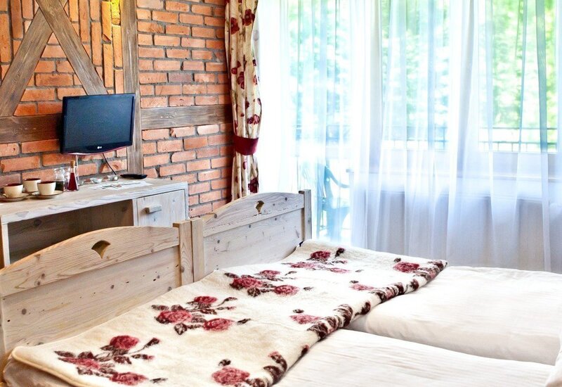 Rustykalne łóżka z kwiatowym kocem, jasne drewniane wezgłowia, ceglany mur z belkami, telewizor, biurko z filiżankami, szyfonowe zasłony i balkon