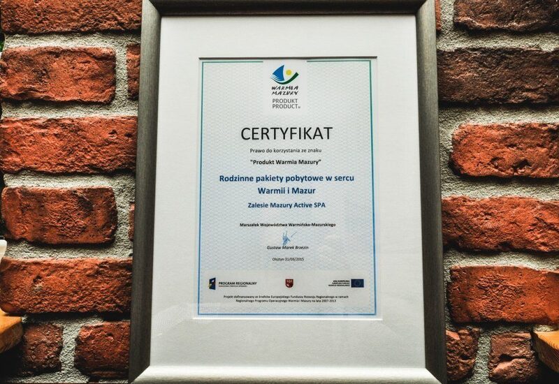Oprawiony certyfikat Zalesie Mazury Active SPA za rodzinne pakiety pobytowe, zawieszony na surowej ceglanej ścianie