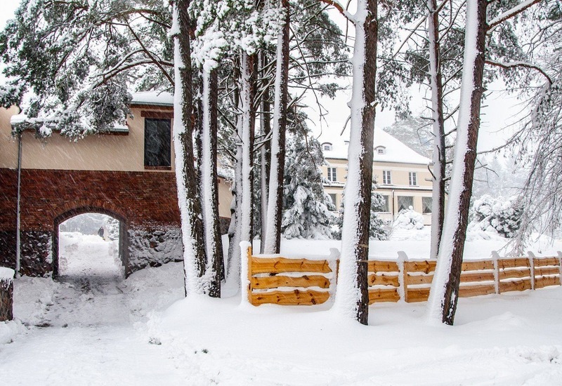 Ośnieżony hotel z arkadowym przejściem, wysokimi sosnami i drewnianym płotem, osoba idzie przez zaśnieżony dziedziniec.