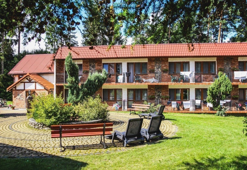 Dwukondygnacyjny pensjonat z czerwonym dachem, balkony z krzesłami i suszącymi ręcznikami, zielone patio z ławkami i leżakami.