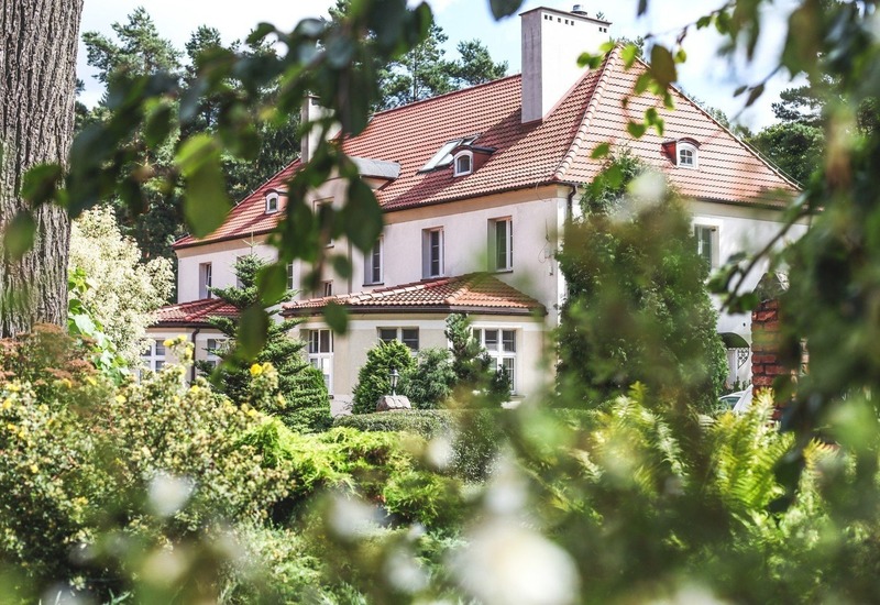 Urokliwa willa z czerwonym dachem, oknami mansardowymi i przeszklonym parterem, otoczona sosnowym lasem i zadbaną zielenią.
