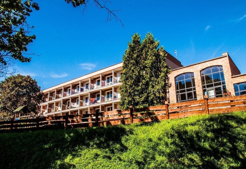 Hotel z rzędami prywatnych balkonów z widokiem na trawiasty stok, drewniany płot, drzewa i duże łukowe okna sali wypoczynkowej