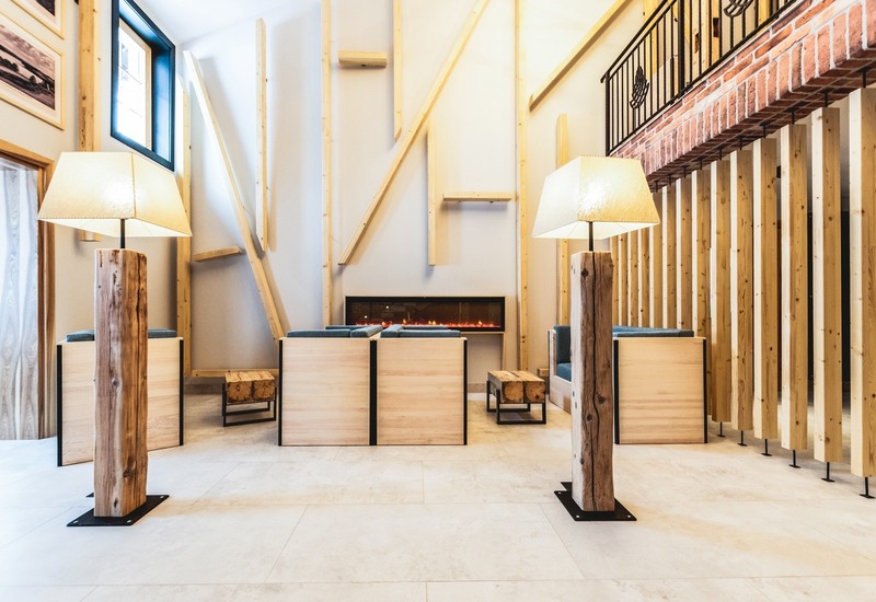 Salon hotelowy w stylu rustykalno-nowoczesnym, lampy na pniach, siedzenia przy długim kominku i antresola z ceglanym wykończeniem