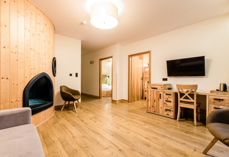 Przestronny apartament z ciepłym drewnianym wykończeniem, zaokrąglonym wbudowanym kącikiem, fotelami, biurkiem z telewizorem i czajnikiem, drzwi do sypialni