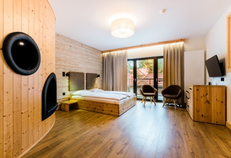 Drewniany pokój hotelowy z niskim łóżkiem na platformie, ciepłym oświetleniem, zaokrągloną ścianą z dwoma czarnymi wnękami, oknem, dwoma fotelami i TV.