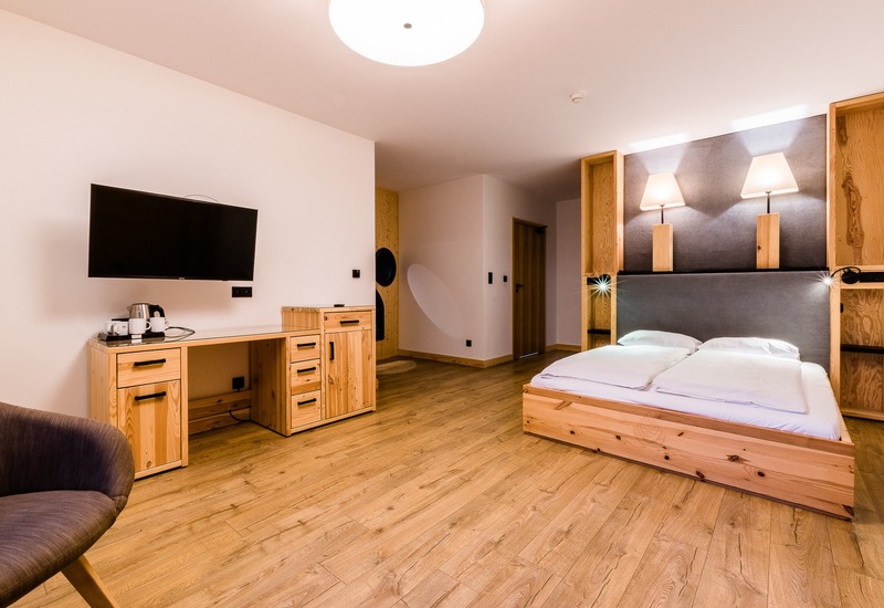 Przestronny pokój hotelowy z drewnianymi meblami, niskim łóżkiem, miękkim zagłówkiem, dwiema lampami, TV i czajnikiem na biurku.