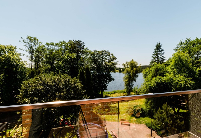 Balkon z przeszkloną balustradą, widok na zieleń, spokojne jezioro i słoneczny taras z ławkami — relaksujący widok nad wodą.