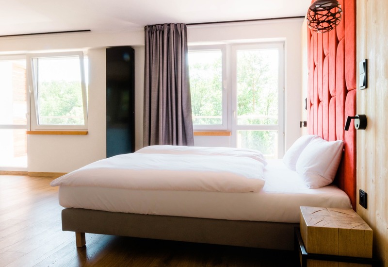 Nowoczesny pokój hotelowy z dużym łóżkiem dwuosobowym, czerwonym pikowanym wezgłowiem, wiszącą lampą, drewnianą podłogą i oknami z widokiem na drzewa.