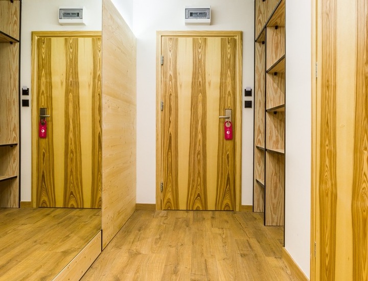 Korytarz hotelowy z ciepłą drewnianą podłogą i drzwiami, lustrem na całą wysokość, otwartymi półkami i czerwonymi zawieszkami nie przeszkadzać