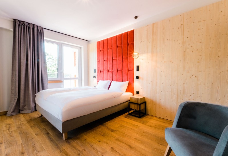 Jasny, nowoczesny pokój hotelowy z czerwonym pikowanym wezgłowiem, dużym łóżkiem, drewnianą ścianą, oknem na balkon i fotelem