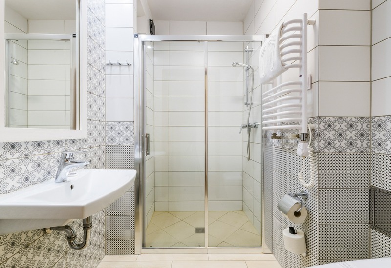 Jasna łazienka hotelowa z przeszklonym prysznicem, umywalką, wzorzystymi płytkami i grzejnikiem na ręczniki z suszarką.