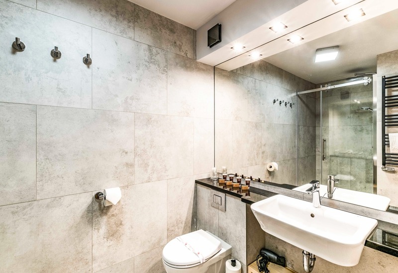 Nowoczesna łazienka hotelowa z szarymi płytkami, przeszklonym prysznicem, umywalką ścienną, kosmetykami na czarnym blacie i grzejnikiem na ręczniki