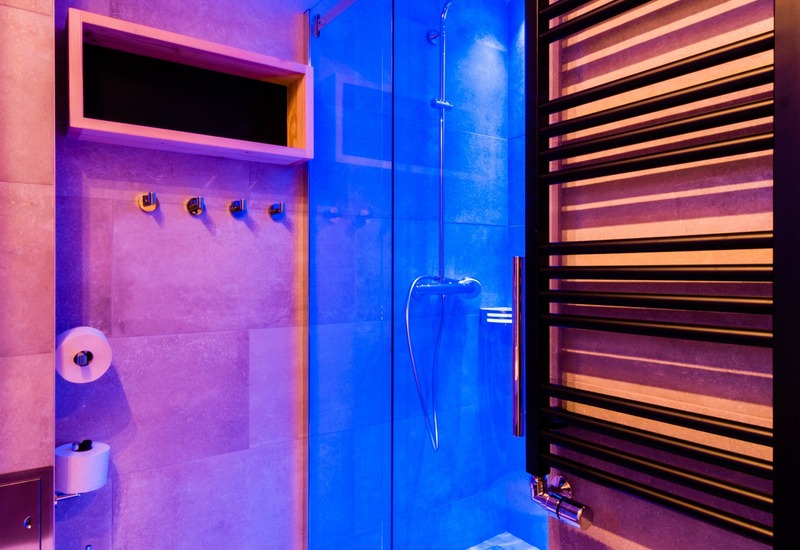 Nowoczesna łazienka hotelowa z przeszklonym prysznicem walk-in i deszczownicą, wiszącą toaletą, grzejnikiem na ręczniki, półką i kolorowym światłem.
