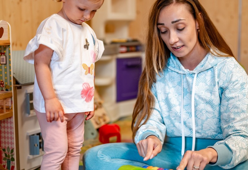 Przytulny kącik zabaw z drewnianymi ścianami — opiekunka bawi się z maluszkiem na kolorowej macie przy kuchni dla dzieci, łodzi i klockach
