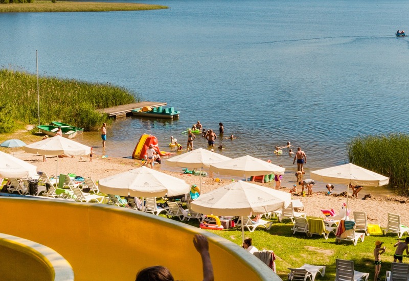 Żółta zjeżdżalnia wodna kończąca się przy piaszczystej plaży nad jeziorem, parasole, leżaki i kąpiące się rodziny.