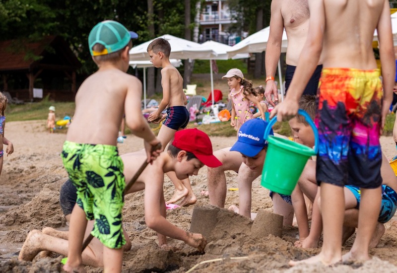 Dzieci budujące zamki z piasku na plaży ośrodka, pod opieką personelu, parasole, leżaki i drzewa w tle.