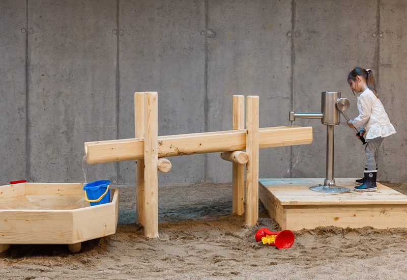 Dziecko kręcące ręczną pompą przy drewnianej instalacji wodnej na wewnętrznym placu zabaw z piaskiem i wiaderkami.
