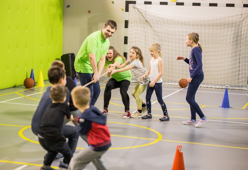 Klub dziecięcy: przeciąganie liny w sali sportowej, opiekunowie w zielonych koszulkach, bramka, pachołki, piłki.