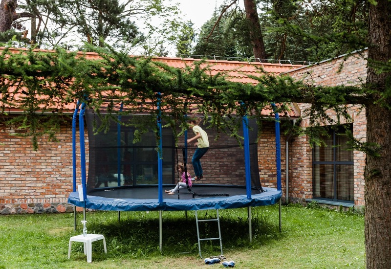 Rodzinny ogród z osłoniętą trampoliną, gdzie dzieci skaczą, na tle ceglanego budynku z czerwonym dachem i wysokimi drzewami.