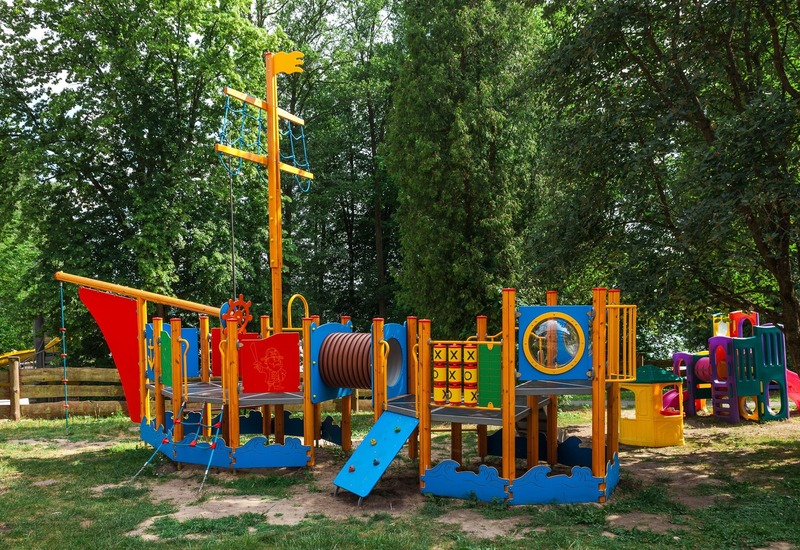 Plac zabaw dla rodzin w kształcie pirackiego statku z zjeżdżalniami, linami i grą kółko-krzyżyk, w cieniu drzew