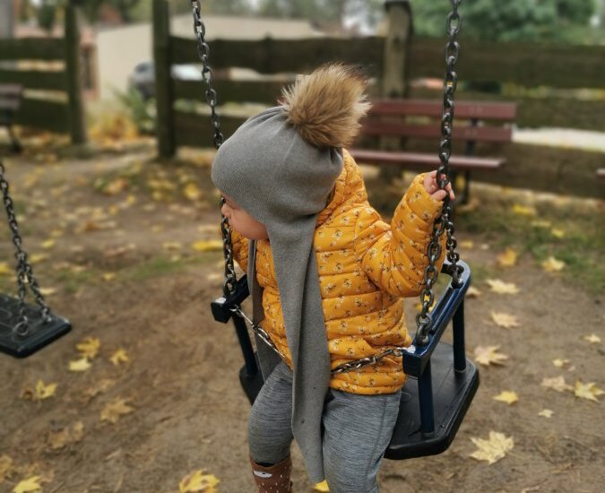 Dziecko w żółtej kurtce i szarej czapce z pomponem na huśtawce w parku, opadłe liście, ławka i drewniany płot