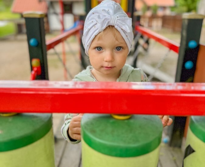 Dziecko w białej chustce z supłem bawi się dużym zielonym ksylofonem na kolorowym placu zabaw, w tle rozmyte domki z czerwonymi dachami