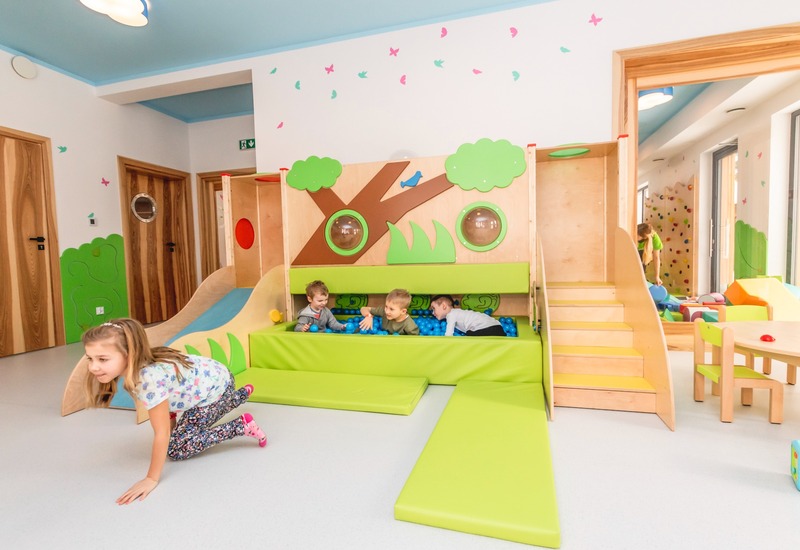 Jasna sala zabaw w hotelu z drewnianą zjeżdżalnią, zielonymi matami, basenem z niebieskich piłek, ścianką wspinaczkową i bawiącymi się dziećmi.
