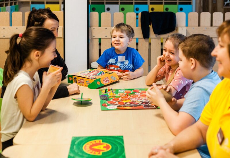 Dzieci grające w gry planszowe przy stoliku klubu dziecięcego, kolorowe pionki i uśmiechnięty opiekun, oferta dla rodzin