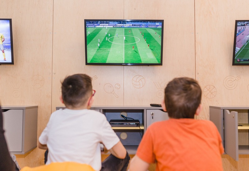 Dwoje dzieci ogląda mecz piłki nożnej na telewizorze w rodzinnej sali gier z konsolą, miejscami do siedzenia i drewnianą ścianą z motywami sportowymi