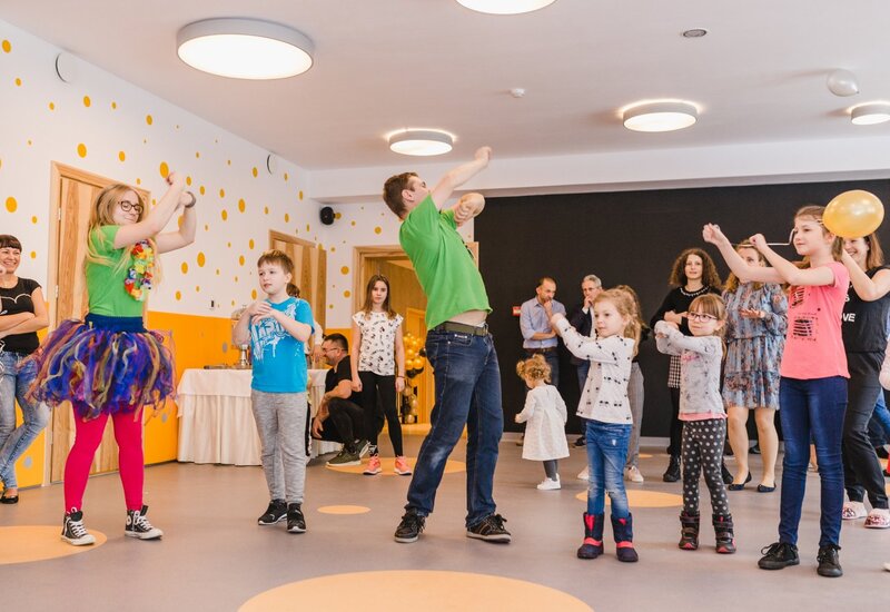 Rodzinna sala eventowa hotelu, dzieci tańczą z animatorką w kolorowej spódnicy, nowoczesne oświetlenie, balony i stolik z poczęstunkiem.