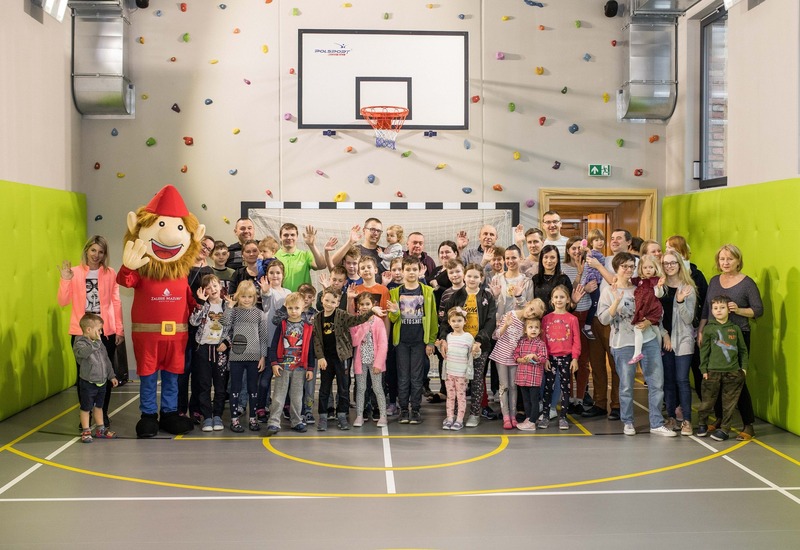 Rodziny i dzieci machające w hotelowej sali zabaw z ścianką wspinaczkową, koszem do koszykówki, bramką i maskotką.