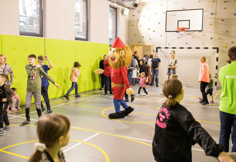 Rodzinna zabawa w sali sportowej z maskotką, ścianką wspinaczkową, koszem do koszykówki i bawiącymi się dziećmi.