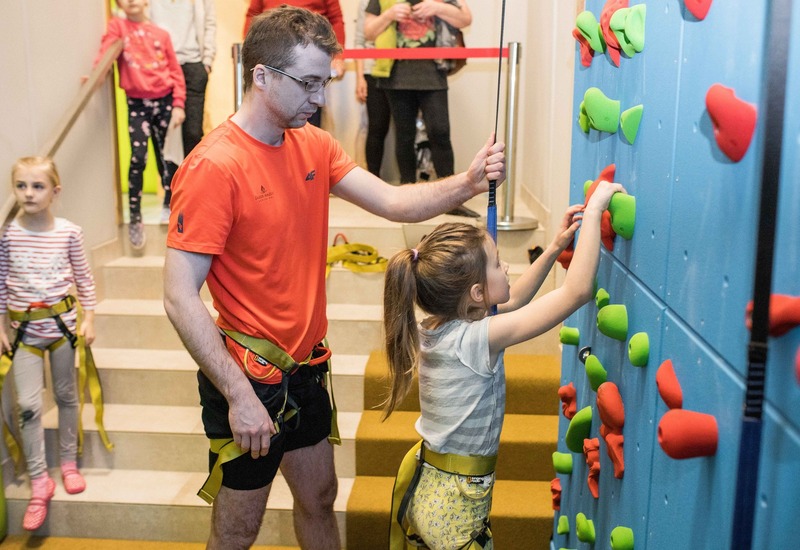 Instruktor pomaga dziewczynce na wewnętrznej ściance wspinaczkowej dla dzieci z kolorowymi chwytami; dzieci w uprzężach czekają na schodach.