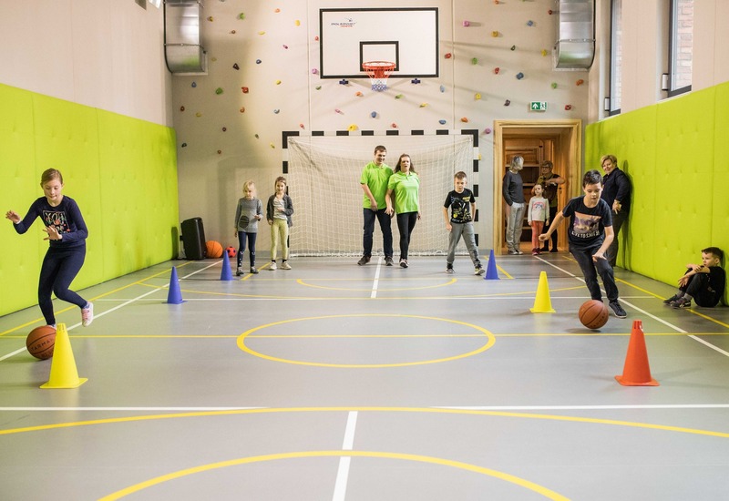 Rodzinna sala sportowa z koszem do koszykówki, ścianką wspinaczkową, jasnozielonymi miękkimi ścianami i pachołkami; dzieci trenują pod opieką.