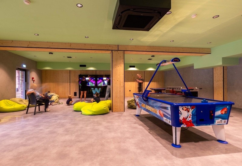 Sala gier z stołem do air hockey, podświetlanym ekranem multiplayer, limonkowymi pufami i drewnianymi belkami; rodziny grają i odpoczywają.