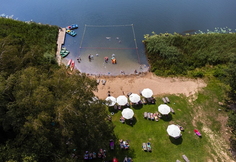 Plaża hotelowa nad jeziorem z wydzielonym kąpieliskiem, drewnianym pomostem i łodziami; trawnik z leżakami, parasolami i rodzinami brodzącymi.